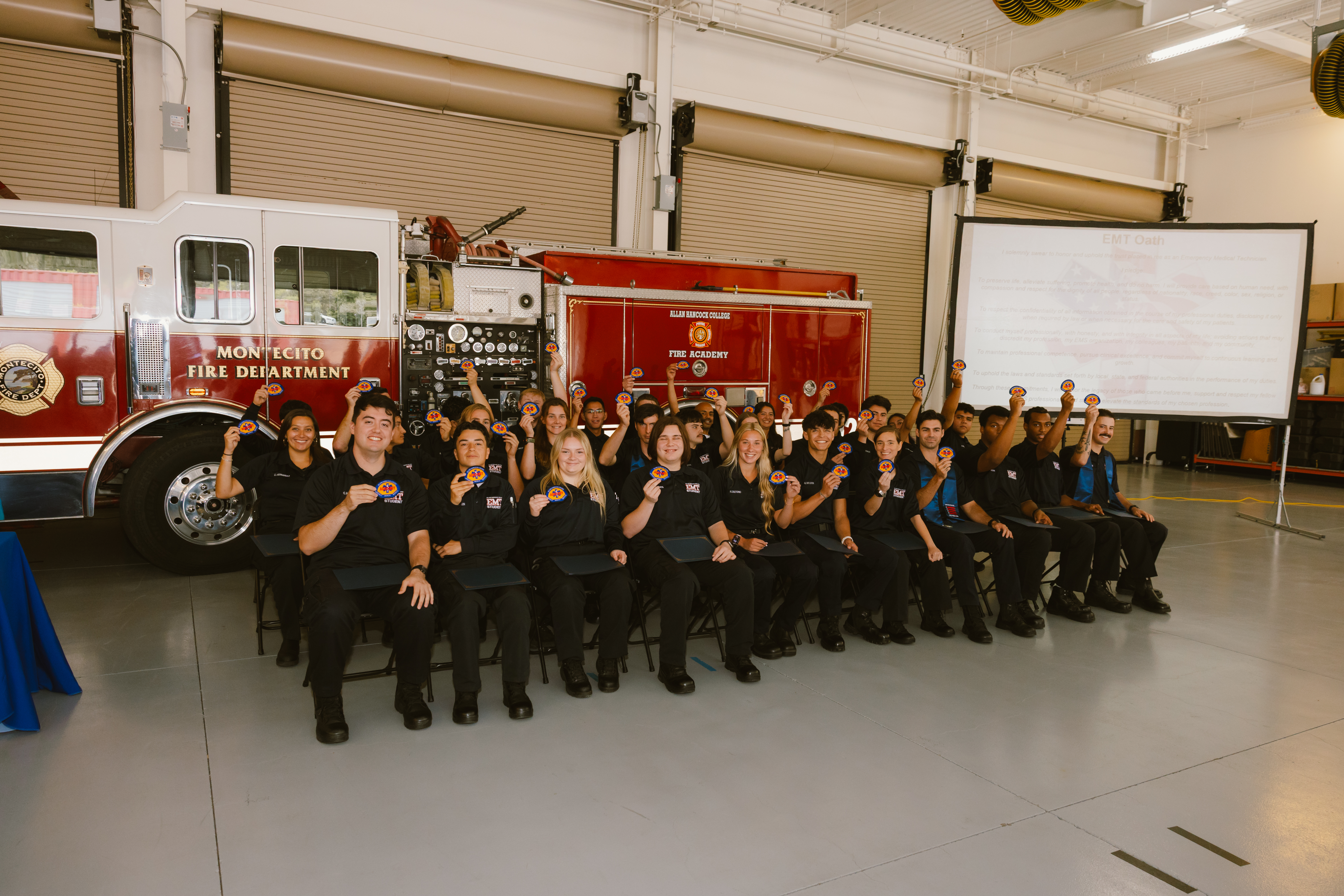 Graduates of Allan Hancock College’s EMS Academy pose for a group photo inside a fire training facility. Wearing black EMS uniforms, the students hold up academy patches and certificates while seated and standing in front of a fire engine. The ceremony marks the completion of their emergency medical services training program